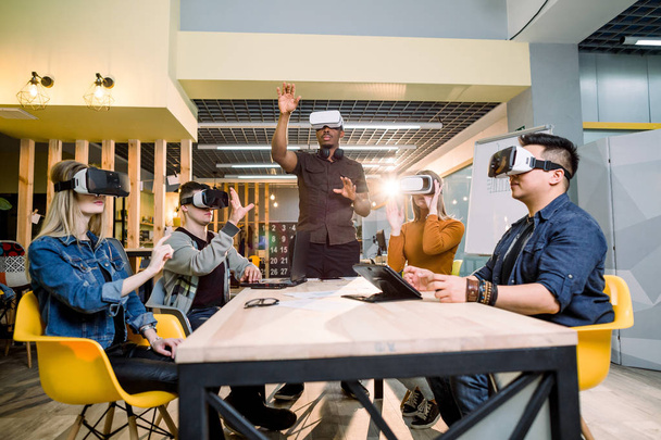 Group of friends and their African man couch trying vr glasses while sitting at the table in office. virtual reality with people having fun together with wearable headset googles - Foto, imagen