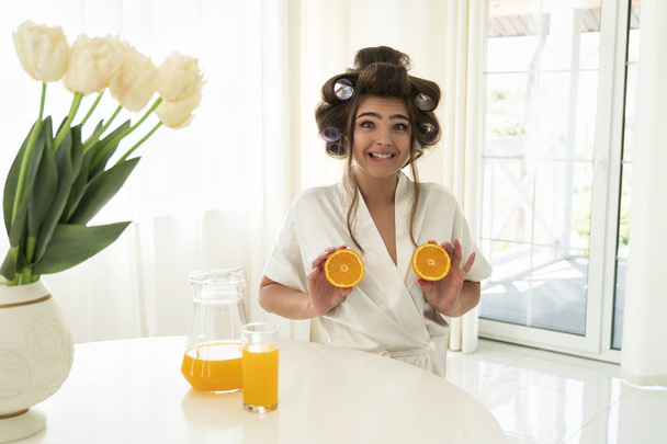 beautiful young brunette woman in hair curlers holding oranges in both hands near her breasts in bright kitchen looking surprised - Fotó, kép