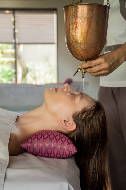 shirodhara, eine ayurvedische Heiltechnik. Öl tropft auf die weibliche Stirn. Porträt einer jungen Frau bei einer ayurvedischen Massage, bei der Aromaöl auf Stirn und Haare tropft. - Foto, Bild