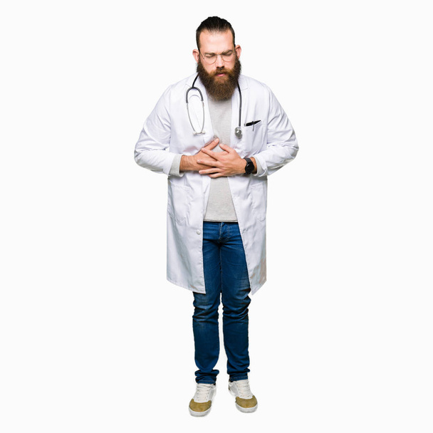 Young blond doctor man with beard wearing medical coat with hand on stomach because indigestion, painful illness feeling unwell. Ache concept. - Photo, Image
