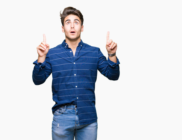 Young handsome man wearing navy shirt over isolated background amazed and surprised looking up and pointing with fingers and raised arms. - Фото, изображение