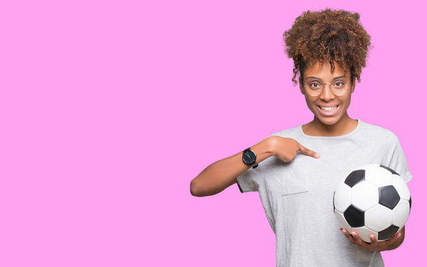 Молодая африканская американка держит футбольный мяч на изолированном фоне с неожиданным лицом, указывающим пальцем на себя
 - Фото, изображение