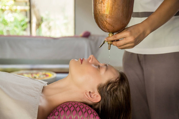 Shirodhara, een Ayurvedische genezings techniek. Olie druipend op het vrouwelijke voorhoofd. Portret van een jonge vrouw tijdens een Ayurvedische massagesessie met aromatische olie druipend op haar voorhoofd en haar. - Foto, afbeelding