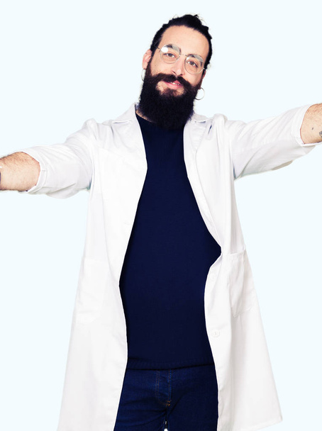 Doctor therapist man with long hair and bear wearing white coat looking at the camera smiling with open arms for hug. Cheerful expression embracing happiness. - Zdjęcie, obraz