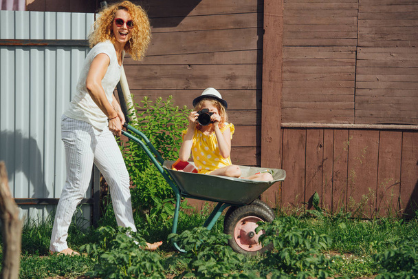 Mother driving a wheelbarrow, while her daughter with camera riding in it - Fotoğraf, Görsel
