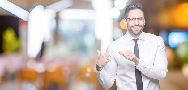 Молодой привлекательный бизнесмен в очках на изолированном фоне указывает на спину с поднятой рукой и большими пальцами, уверенно улыбаясь
 - Фото, изображение