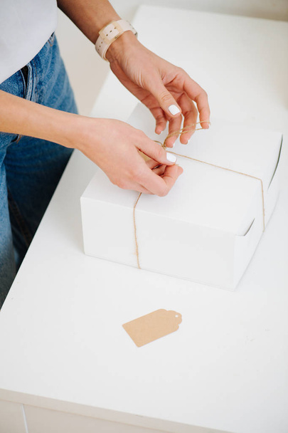 Woman in casual clothes is tying postal cardboard box, attaching tag at home - 写真・画像
