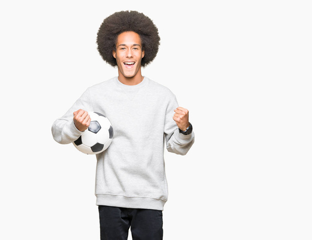Jovem americano africano com cabelo afro segurando bola de futebol gritando orgulhoso e celebrando vitória e sucesso muito animado, animando emoção
 - Foto, Imagem