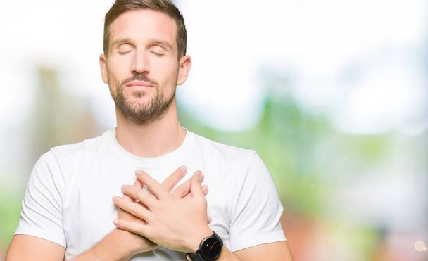 Handsome man wearing casual white t-shirt smiling with hands on chest with closed eyes and grateful gesture on face. Health concept. - 写真・画像