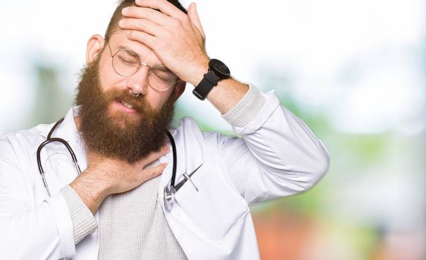 Fiatal szőke orvos férfi szakáll viselése, orvosi kabát megható a betegség és láz, influenza és nátha, vírus beteg homlokára - Fotó, kép
