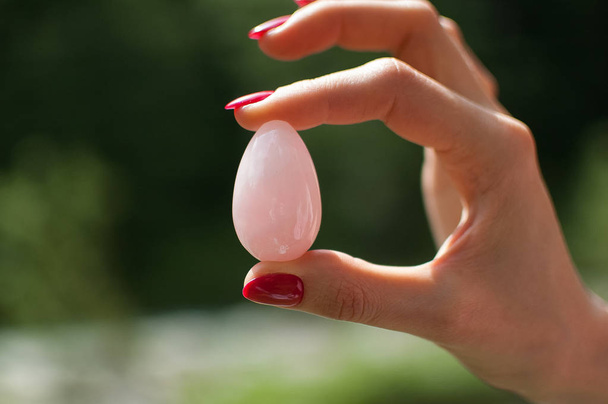 Κλείστε τη φωτογραφία του γυναικείου χεριού κρατώντας ένα αυγό Γιόνι. Κρύσταλλο Ροζέ χαλαζία στο φόντο του ποταμού. Ιδέα για την υγεία των γυναικών - Φωτογραφία, εικόνα