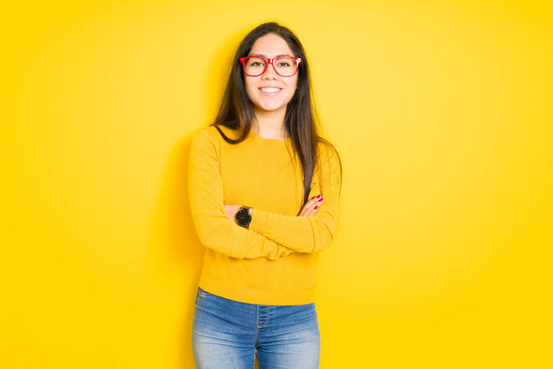 Beautiful brunette woman wearing red glasses over yellow isolated background happy face smiling with crossed arms looking at the camera. Positive person. - Фото, изображение