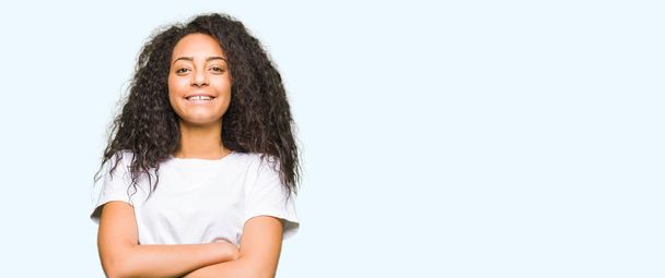 Krásná mladá dívka s kudrnatými vlasy nosí ležérní bílé tričko Veselý obličej s úsměvem překřížené paže při pohledu na fotoaparát. Pozitivní člověk. - Fotografie, Obrázek