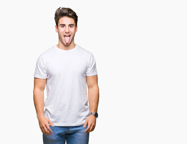junger gutaussehender Mann in weißem T-Shirt über isoliertem Hintergrund, der die Zunge mit lustigem Gesichtsausdruck herausstreckt. Emotionales Konzept. - Foto, Bild