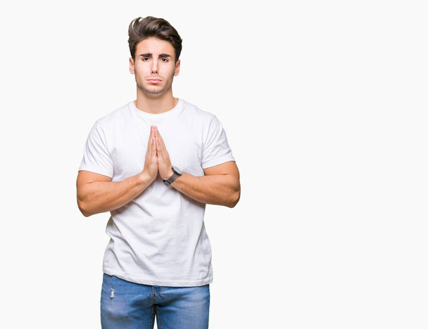 Young handsome man wearing white t-shirt over isolated background begging and praying with hands together with hope expression on face very emotional and worried. Asking for forgiveness. Religion concept. - Foto, immagini