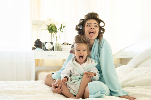 beautiful young brunette woman and cute baby girl wearing blue bathrobes with curlers having fun on the bed in bright bedroom - Foto, Bild