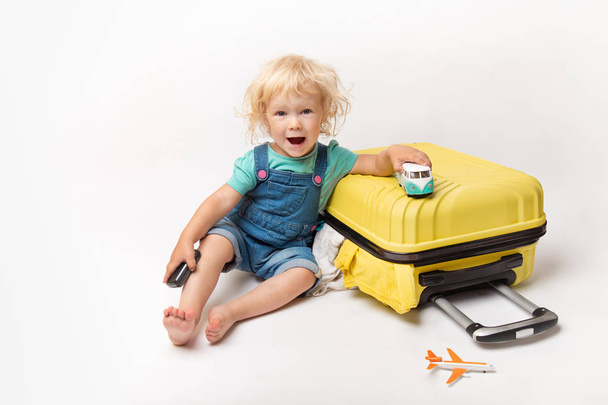 Little Happy Caucasian curly girl in comfortable denim clothes child sitting on yellow suitcase. tourist in shirt with plain And car in hand. Orange suitcase. Summer vacation. - Φωτογραφία, εικόνα