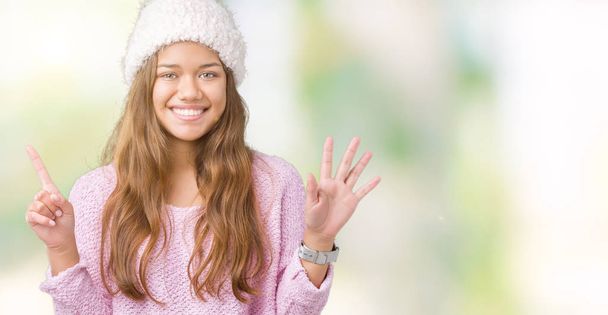 Numara altı ise kendinden emin ve mutlu gülümseyen genç güzel esmer kadın giyiyor kazak ve kış şapka gösterilen ve parmakları ile işaret izole arka plan üzerinde. - Fotoğraf, Görsel