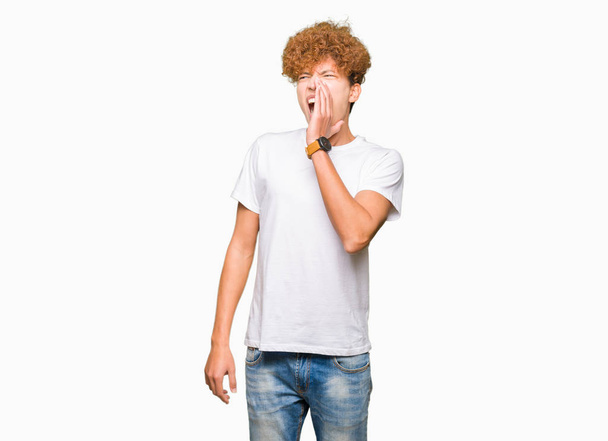 junger gutaussehender Mann mit Afro-Haaren, lässig in weißem T-Shirt schreiend und laut schreiend mit der Hand auf dem Mund. Kommunikationskonzept. - Foto, Bild