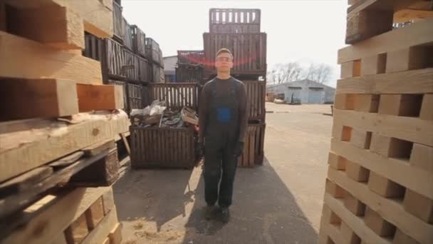 Un joven trabajador en uniforme camina entre paletas de madera en almacén de distribución al aire libre. Movimiento lento
 - Metraje, vídeo
