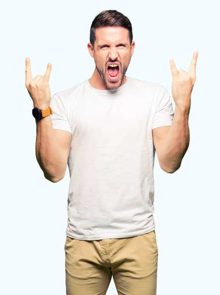 Pohledný muž nosí ležérní bílé tričko křik s šílený výraz dělá rock symbol s rukama. Hudební hvězda. Těžký koncepce. - Fotografie, Obrázek