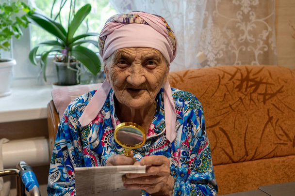 Zeer oude vrouw met vergrootglas die probeert te lezen uit een krant. grootmoeder 90 jaar oud leest aan de tafel met vergrootglas. Een close-up portret - Foto, afbeelding