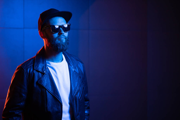 Hipster handsome man on the city streets being illuminated by neon signs. He is wearing leather biker jacket or asymmetric zip jacket with black cap, jeans and sunglasses. - Foto, Imagem