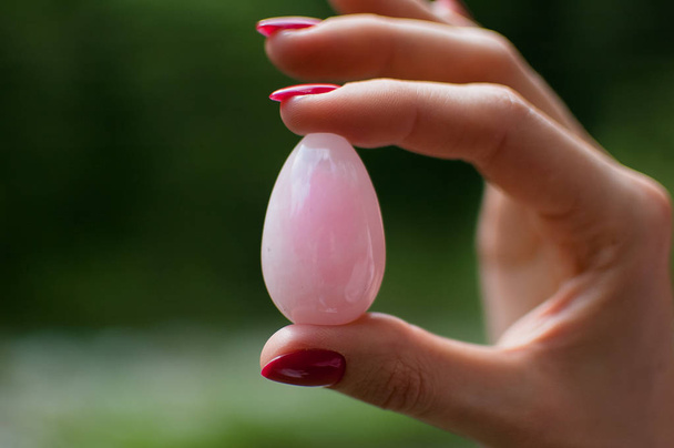 Foto de cerca de la mano femenina sosteniendo un huevo de yoni. Huevo de cristal de cuarzo rosa sobre fondo de río. Concepto de salud para mujeres
 - Foto, imagen