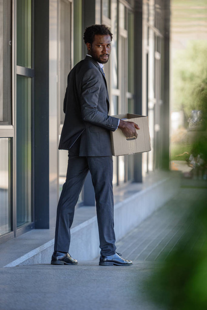 enfoque selectivo del empresario afroamericano despedido mirando hacia atrás mientras sostiene la caja de cartón
 - Foto, imagen