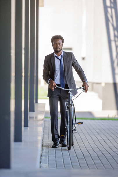 pensativo hombre de negocios afroamericano caminando cerca de edificio de oficinas con bicicleta
 - Foto, Imagen