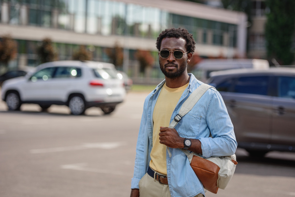όμορφος Αφρικανός Αμερικανός άνθρωπος στα γυαλιά ηλίου κοιτάζοντας την κάμερα, ενώ στέκεται στο πάρκινγκ του αυτοκινήτου - Φωτογραφία, εικόνα