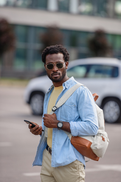Afroameričan, který drží telefon a dívá se na kameru, když stojí na parkovišti - Fotografie, Obrázek