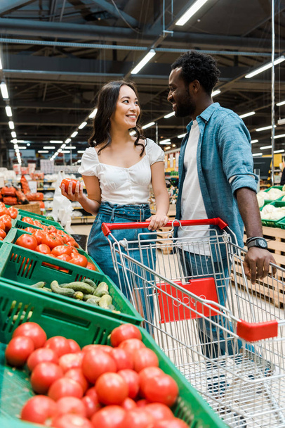 selektiver Fokus glücklicher asiatischer Mädchen und afrikanisch-amerikanischer Männer, die neben frischen Tomaten im Supermarkt stehen  - Foto, Bild