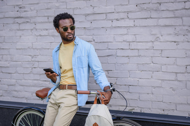 stylischer afrikanisch-amerikanischer Geschäftsmann mit Sonnenbrille, der mit Fahrrad in der Nähe von Ziegelmauer steht und Smartphone in der Hand hält - Foto, Bild