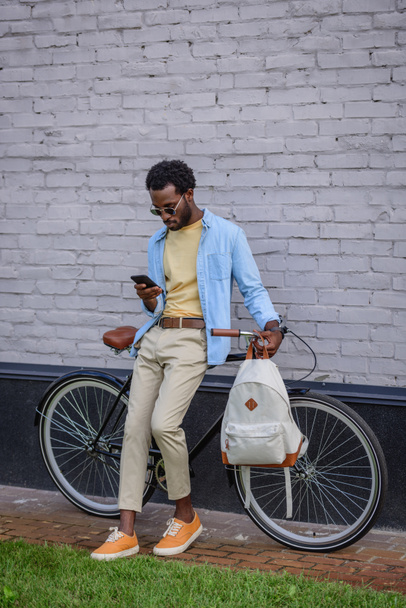 handsome african american man using smartphone while standing with bike near brick wall - 写真・画像