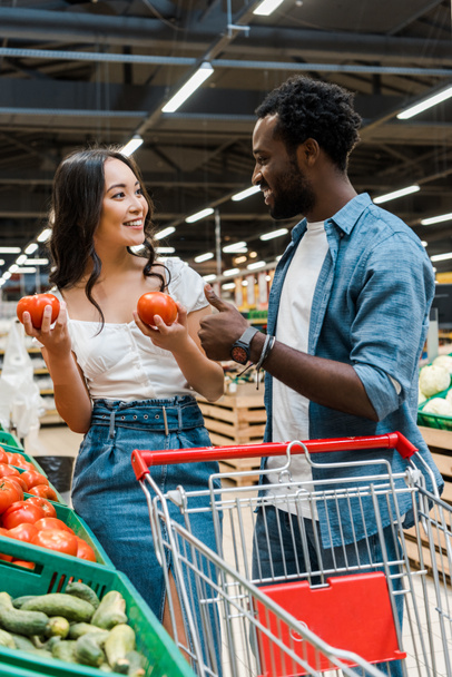избирательный фокус счастливой азиатской женщины, держащей свежие помидоры рядом с веселым африканским американским мужчиной, показывающим большой палец вверх в супермаркете
  - Фото, изображение