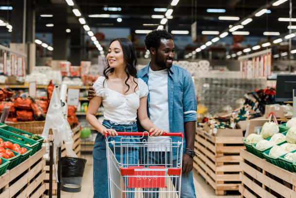 陽気なアフリカ系アメリカ人男性とスーパーマーケットのショッピングカートの近くに幸せなアジアの女性  - 写真・画像