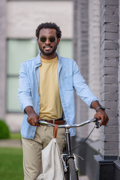 Stylowy African American Man w okulary patrząc na kamerę stojąc w pobliżu roweru - Zdjęcie, obraz