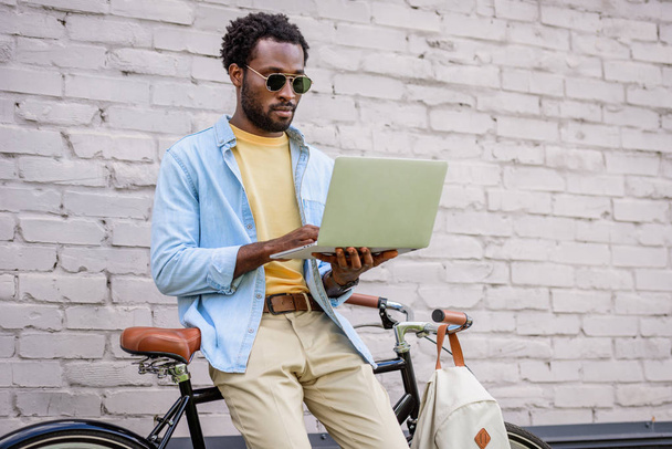 uomo afroamericano attento ed elegante utilizzando il computer portatile mentre in piedi vicino al muro di mattoni vicino alla bicicletta
 - Foto, immagini