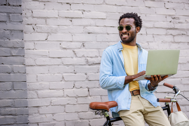 glimlachend African American man op zoek weg terwijl staande door bakstenen muur in de buurt van fiets en het gebruik van laptop - Foto, afbeelding
