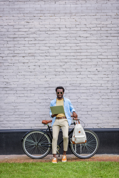 hombre afroamericano elegante de pie junto a la pared de ladrillo cerca de la bicicleta y el uso de teléfono inteligente
 - Foto, Imagen