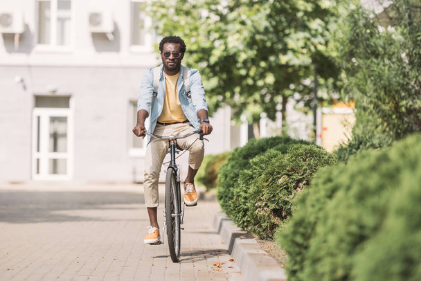 knappe Afrikaanse Amerikaanse man paardrijden fiets langs Sunny Street en groene planten - Foto, afbeelding
