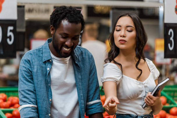 atractivo asiático mujer con notebook apuntando con dedo cerca feliz africano americano hombre
  - Foto, Imagen