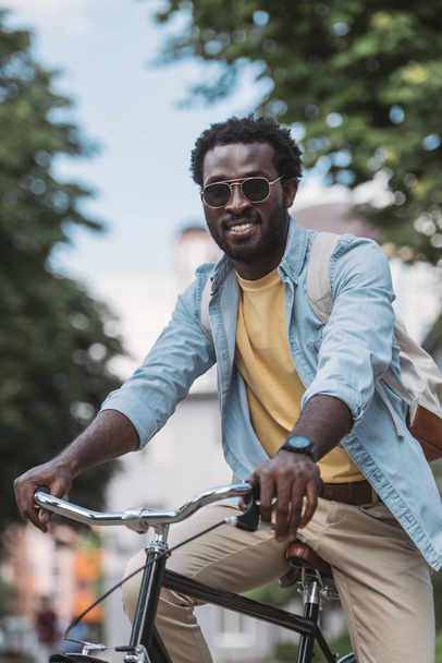 веселий афроамериканський чоловік в сонцезахисних окулярах посміхається на камеру під час їзди на велосипеді
 - Фото, зображення