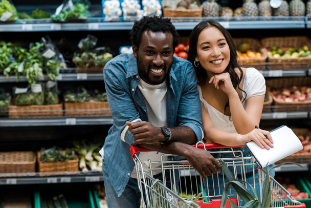 heureux couple interracial souriant dans supermarché près du panier
  - Photo, image