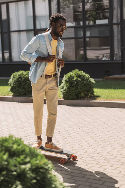 handsome, stylish african american man smiling while riding longboard on sunny street - Foto, afbeelding