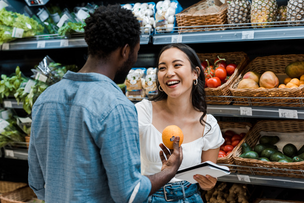  cheerful asian girl near african american man with orange in store - Photo, Image