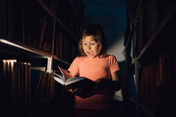 malé dítě čte knihu, je překvapen, má otevřenou pusu. drží v ruce knihu, učí se v knihovně, v interiéru 1 - Fotografie, Obrázek