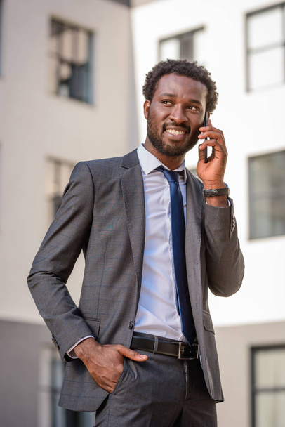cheerful african american man holding hand in pocket and looking away while talking on smartphone - Foto, afbeelding