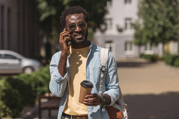 stylish african american man in sunglasses talking on smartphone and holding paper cup - Foto, afbeelding